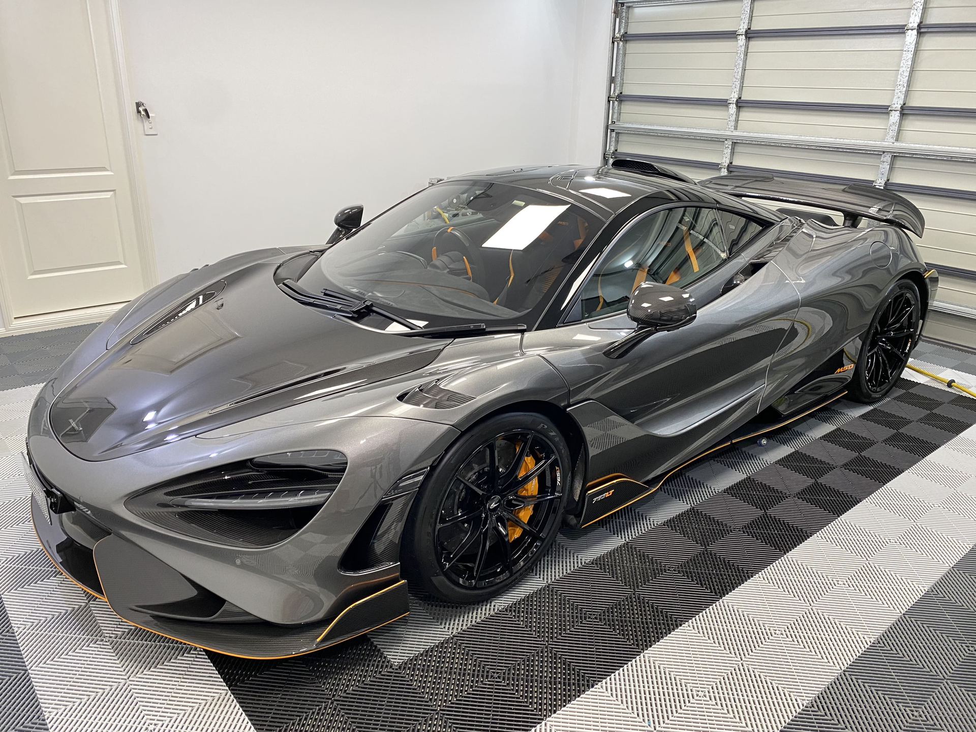A dark grey McLaren sports car parked in a garage on a black and white tiled floor.