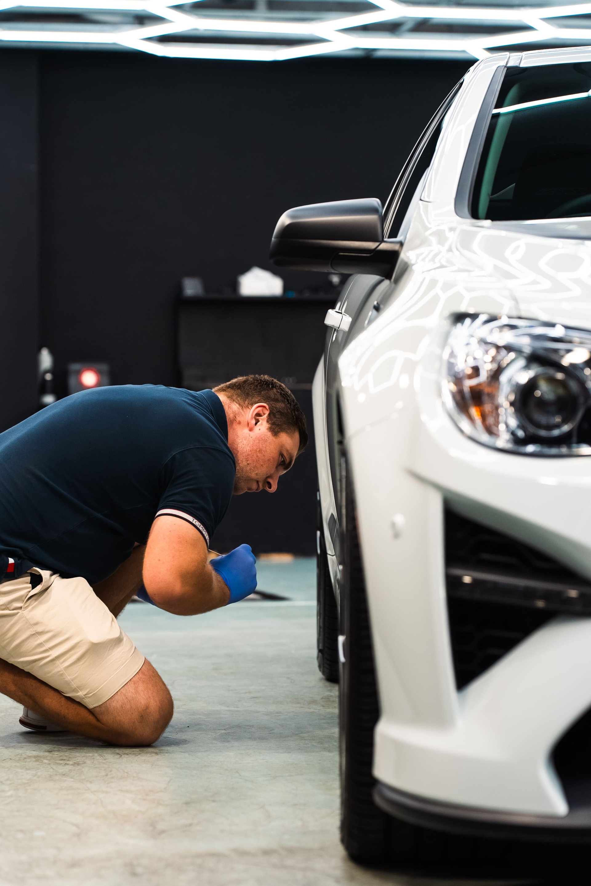 Man in blue gloves examining the front tire of a white car indoors.