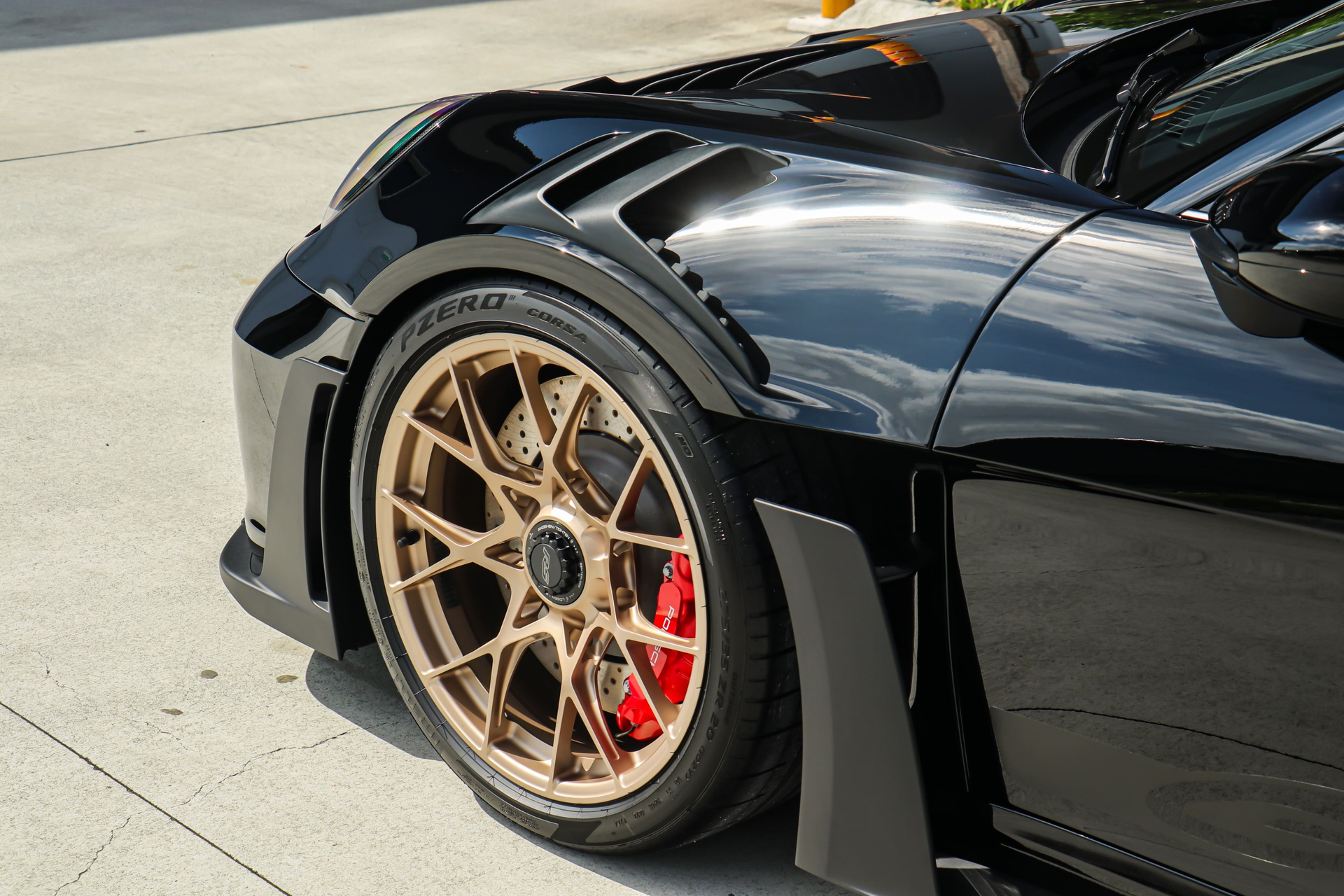 A close-up view of the front wheel and fender of a black sports car with bronze wheels and red brake calipers.