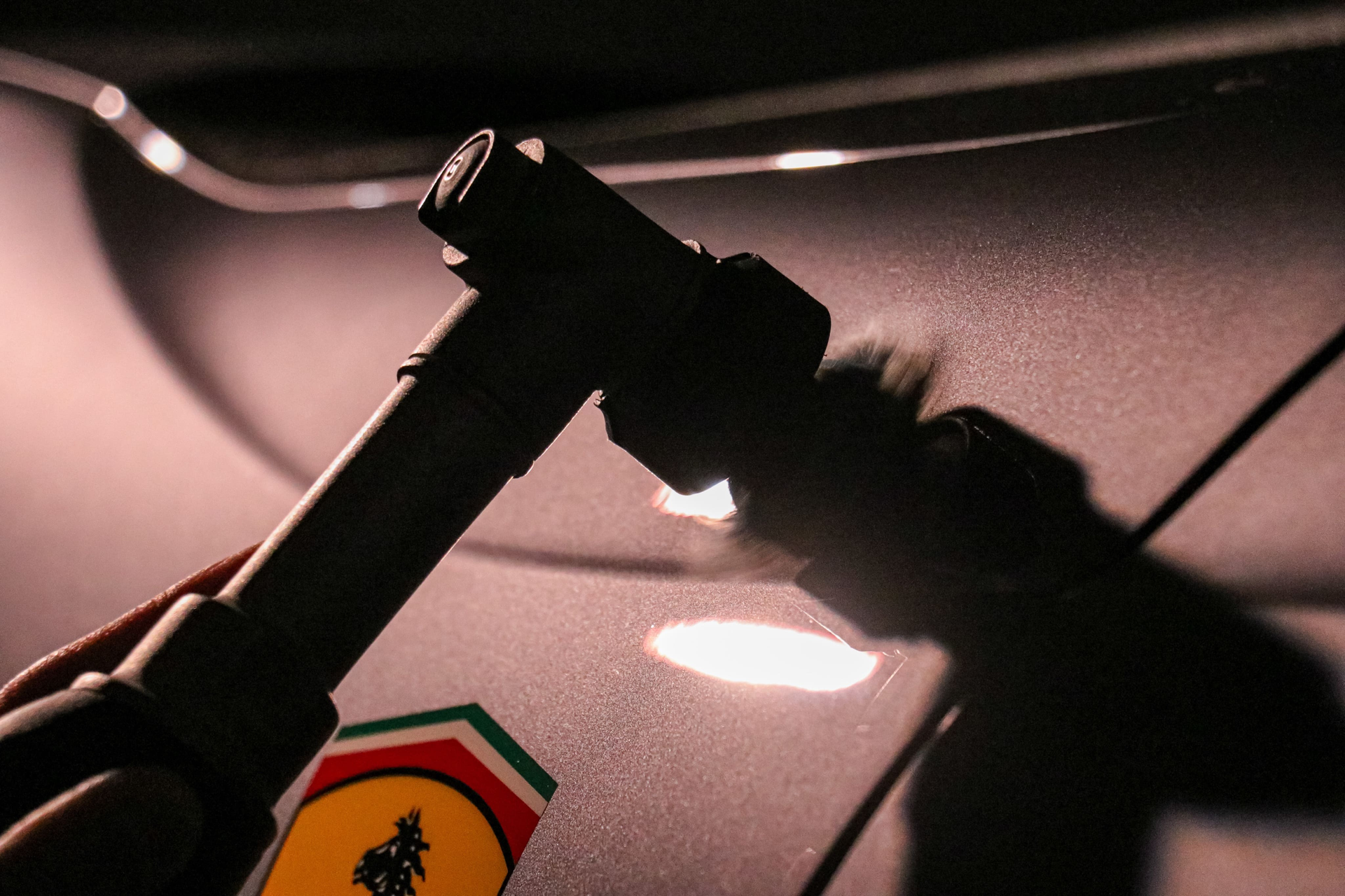 Close-up of a car's surface with a dark object held up, casting a shadow. A Ferrari logo is visible.
