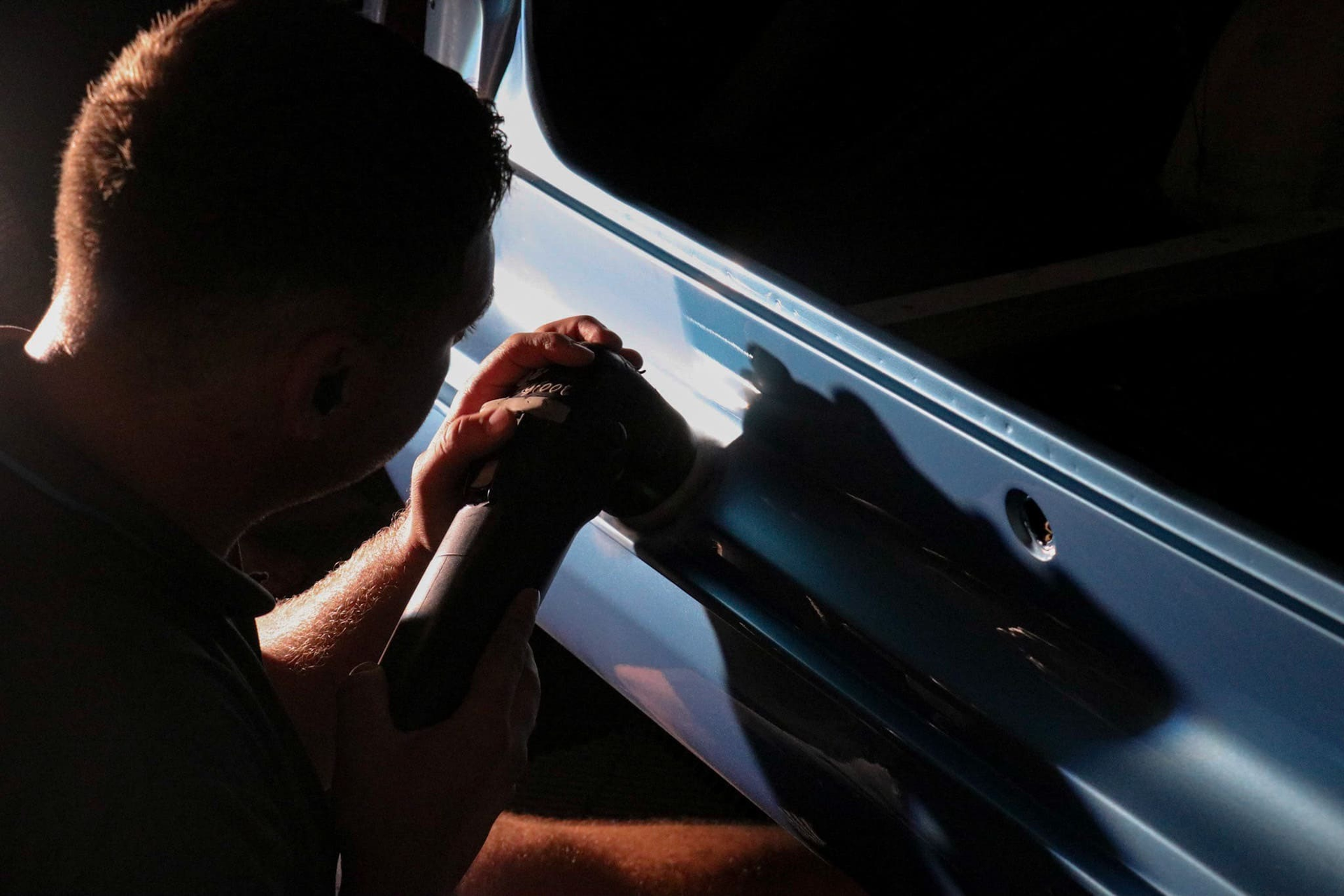 A person uses a handheld power polisher to buff the side panel of a blue car in a dimly lit setting.