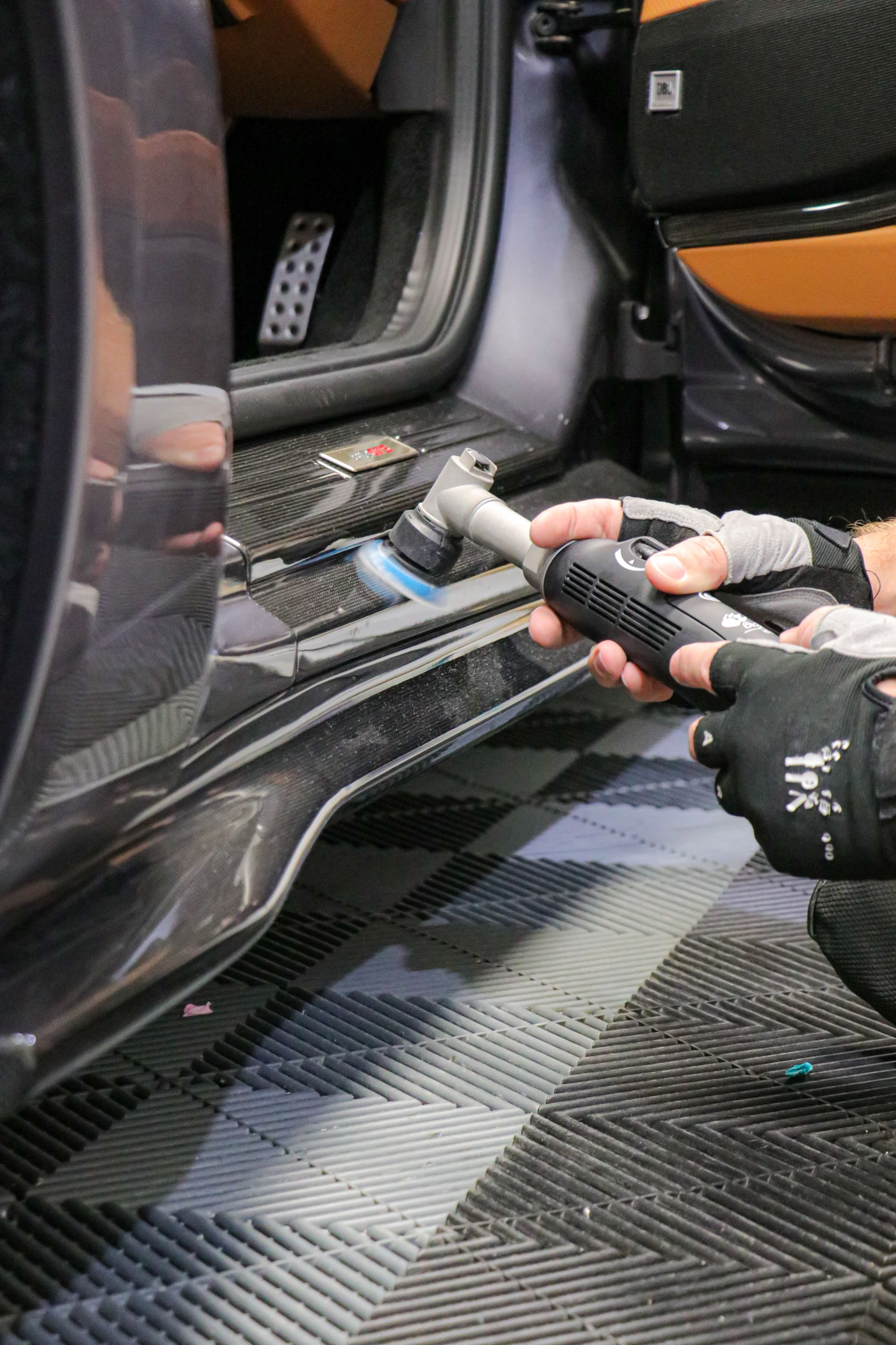 A person using a power polisher on a car's side skirt; in a garage setting with a checkered floor.
