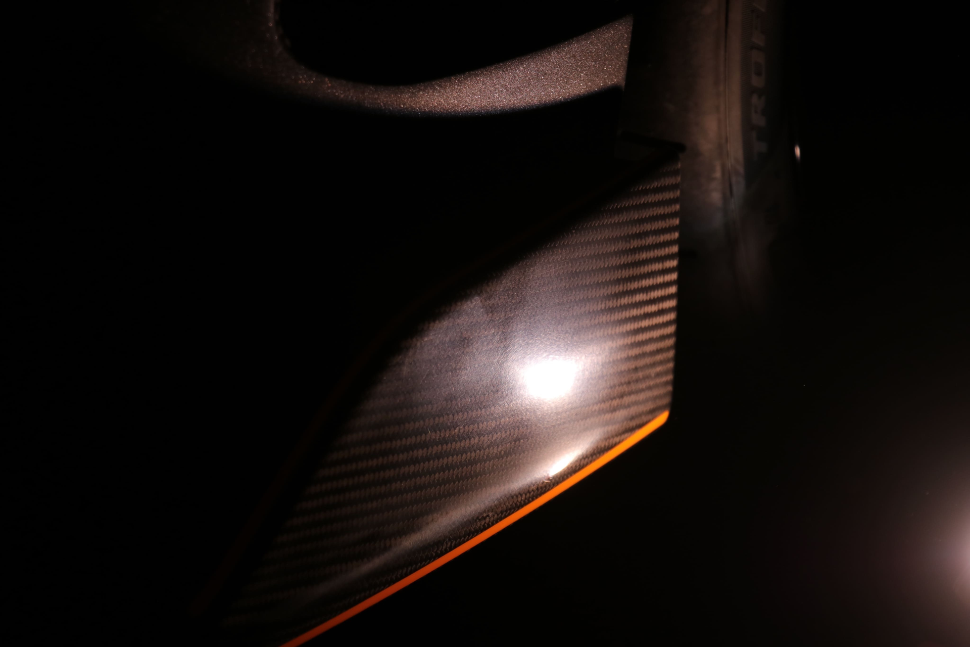Close-up of a carbon fiber surface with a visible diagonal weave pattern, illuminated by a bright, central light reflection.