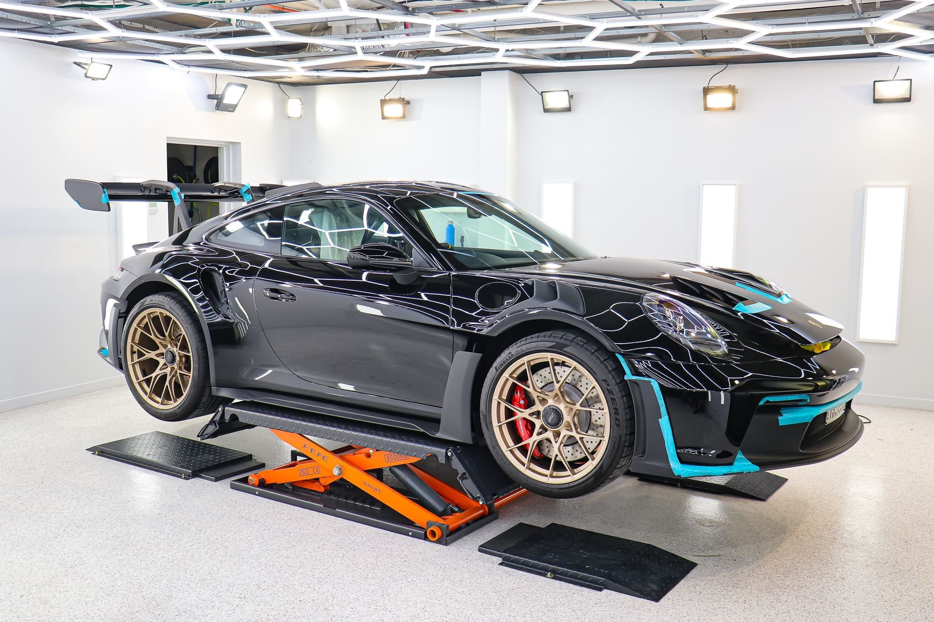 A black Porsche sports car with teal accents and bronze wheels raised on an orange lift in a modern, well-lit studio.