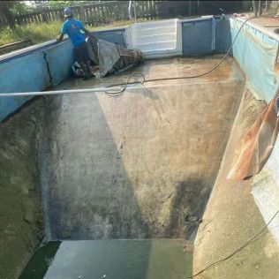 Man cleaning a drained, dirty pool. Concrete pool has light blue trim, white steps, and a bit of water remains.