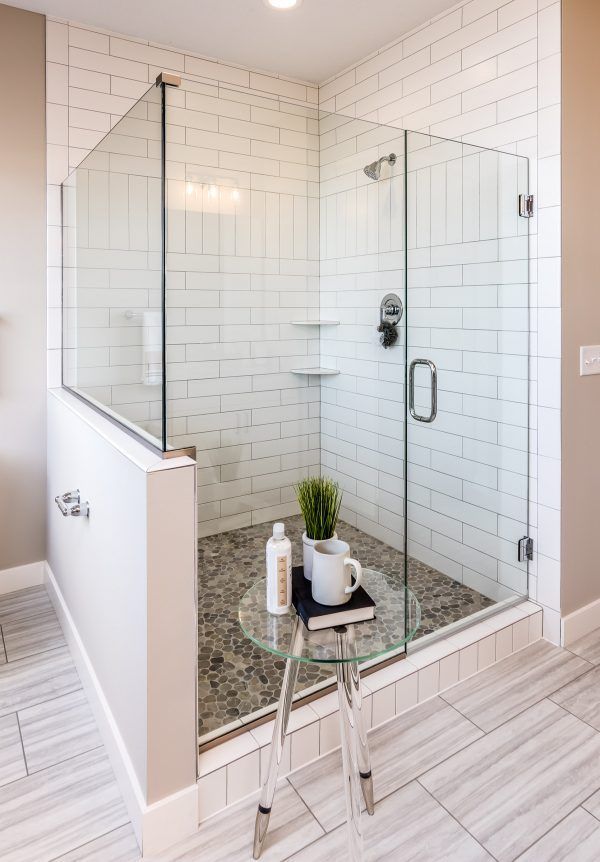 A modern bathroom with a glass shower enclosure, white subway tiles, and stone floor. A glass table holds bath items.