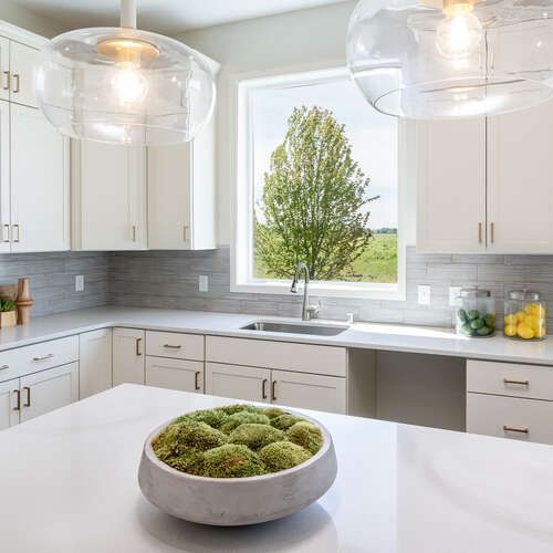 Modern white kitchen with island, large window with tree view, and orb pendant lights.