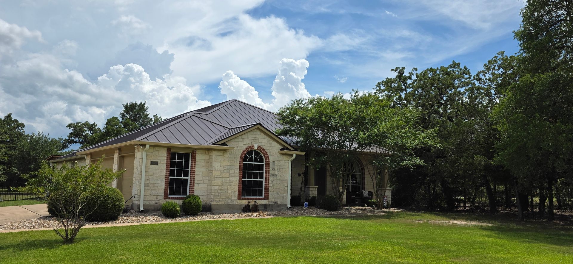 A single-story house with stone siding and standing seam roof under a cloudy sky, surrounded by green grass and trees.