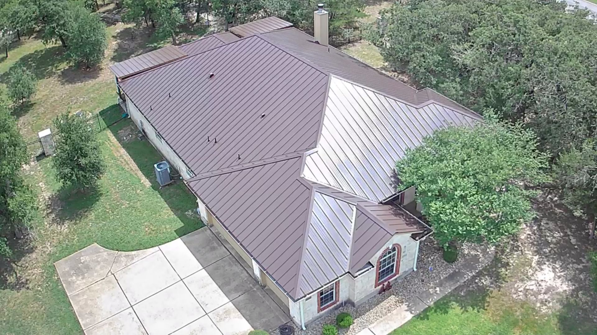Maroon metal roof on a house, aerial view. Trees surround a concrete driveway and lawn.