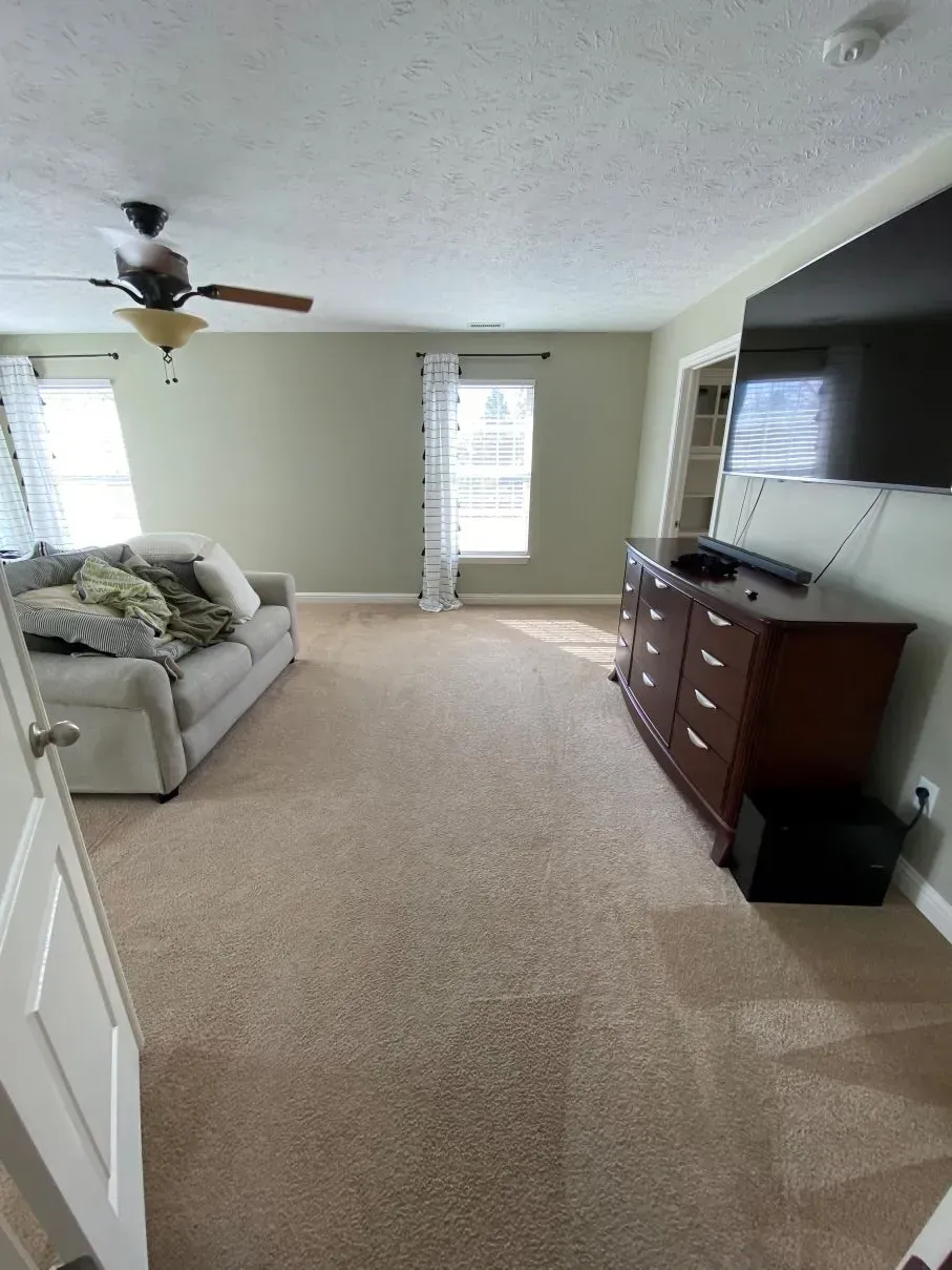 Spacious room with a sofa, dresser, TV, window, and ceiling fan; beige carpet and walls.