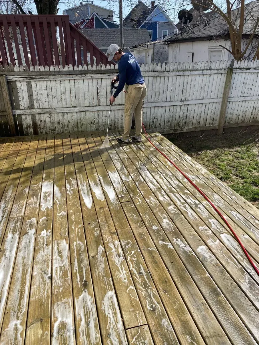 Person power washing a wooden deck, removing dirt. White fence and houses in the background.