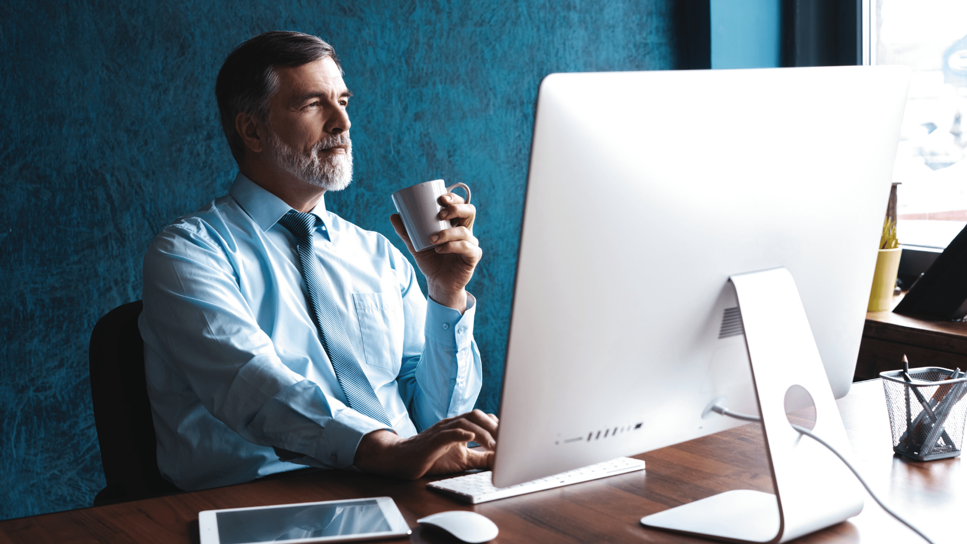 A man is sitting at a desk in front of a computer holding a cup of coffee.