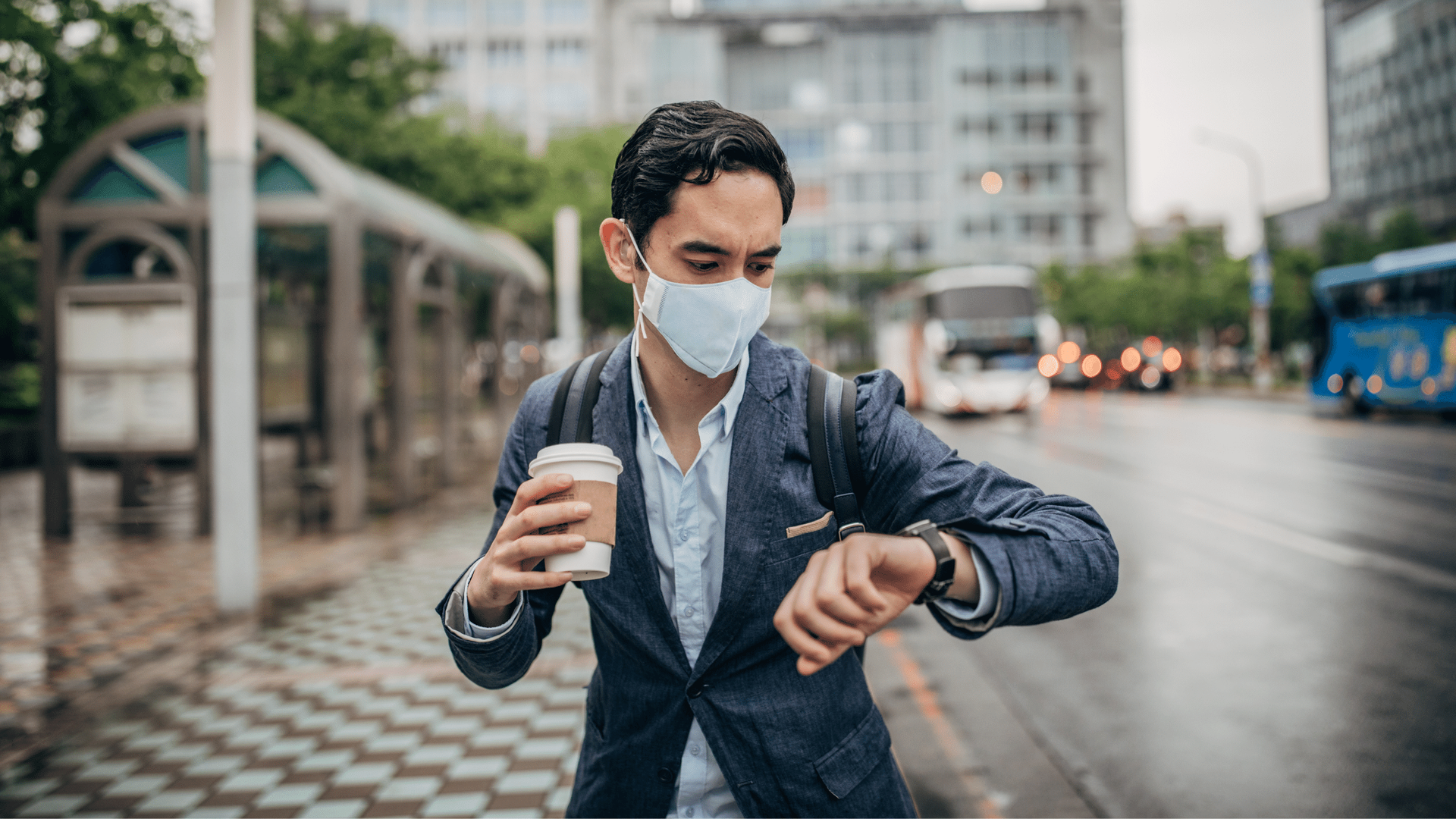 A man wearing a mask is looking at his watch while holding a cup of coffee.