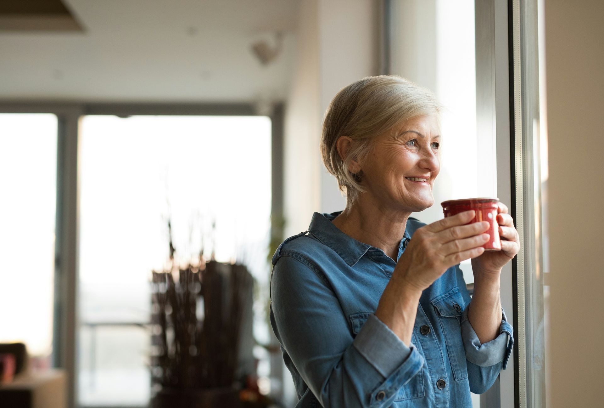 An elderly woman is drinking a cup of coffee and looking out of a window.