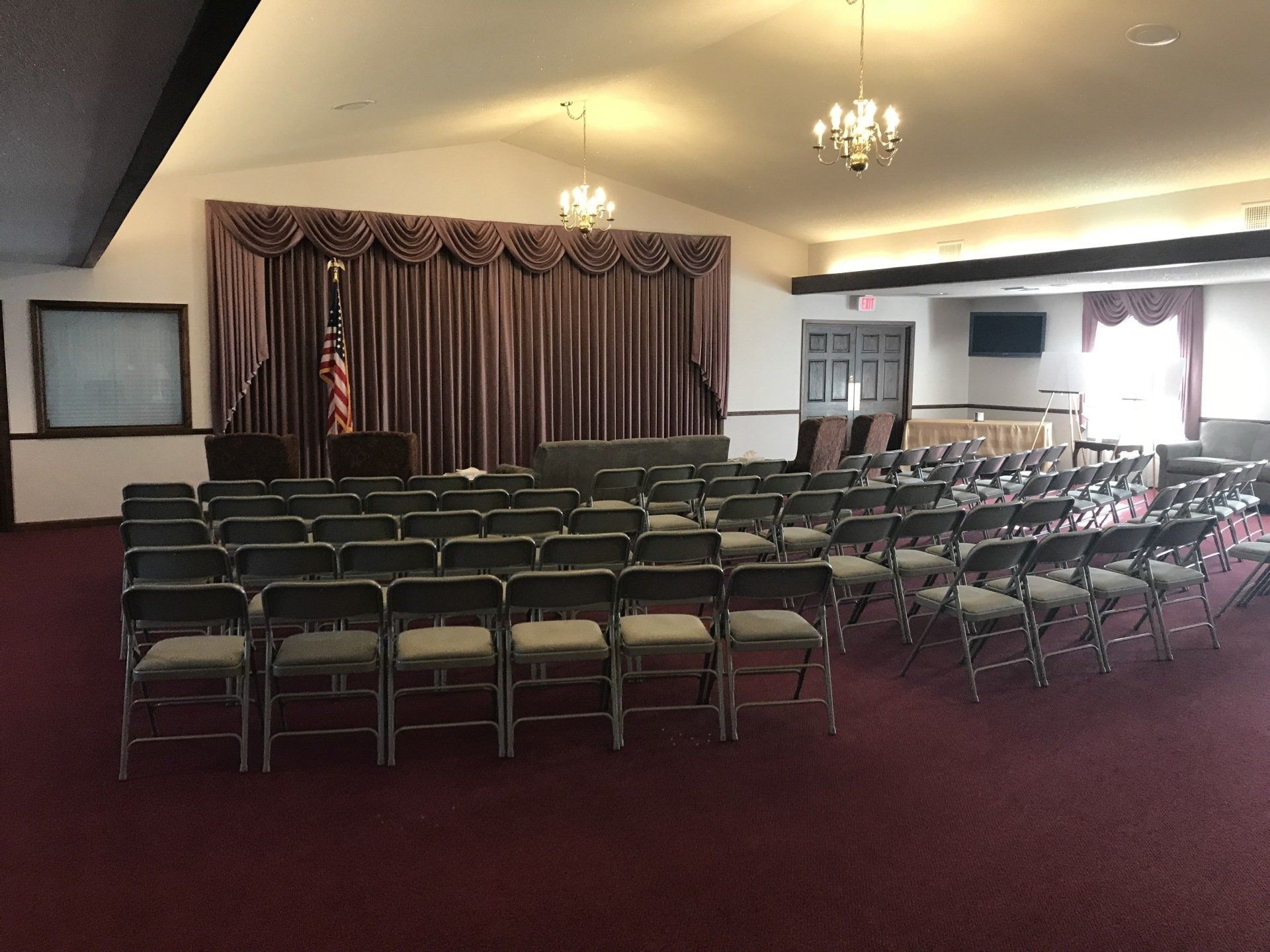 Rows of empty chairs face a stage with maroon curtains and an American flag in a carpeted room.