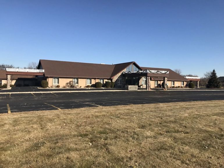 Brown-roofed brick building with a large parking lot, set against a blue sky, and dry grassy field in front.