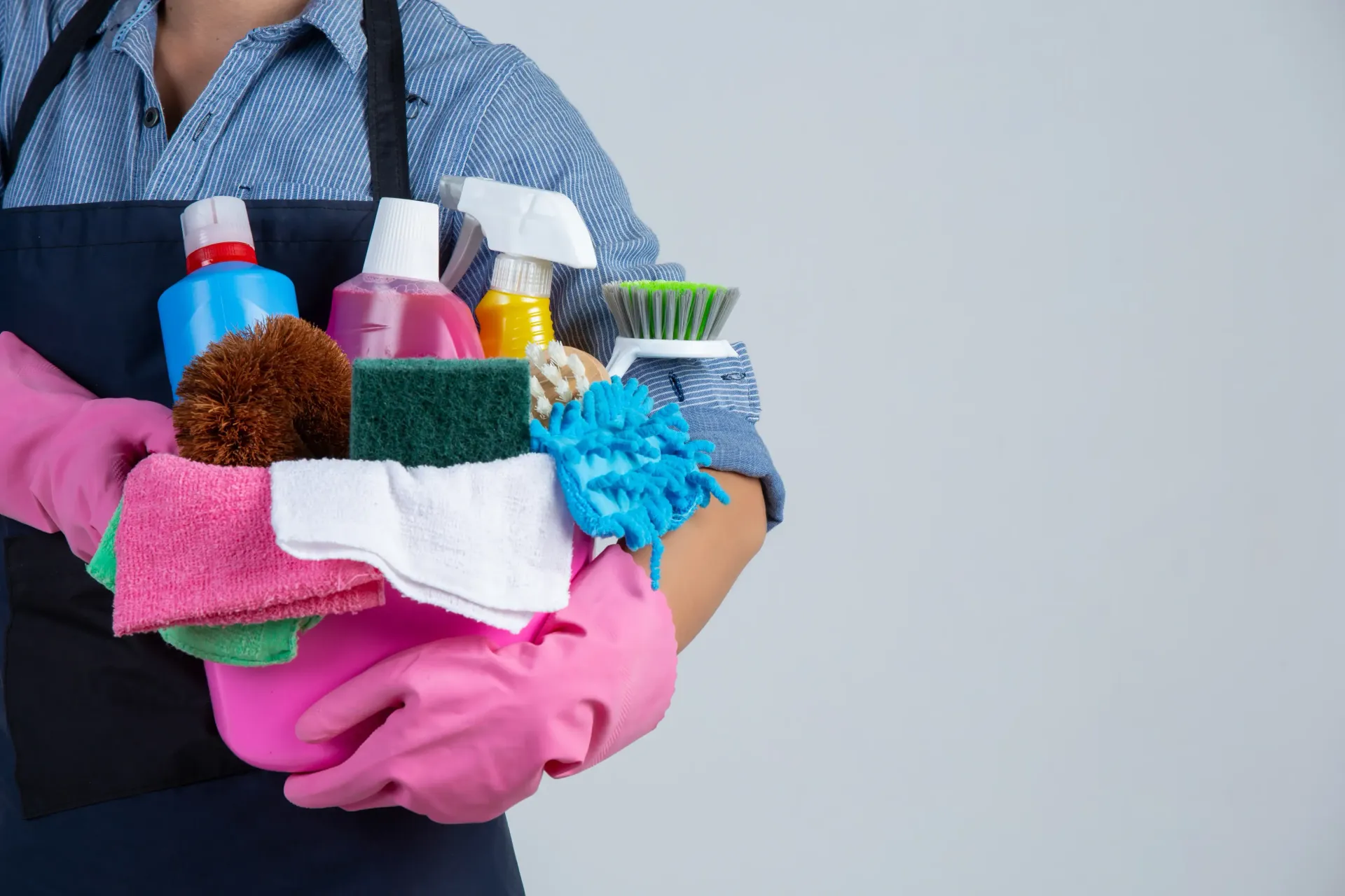 Person in apron holding cleaning supplies, including bottles, brushes, and cloths. Wearing pink gloves.