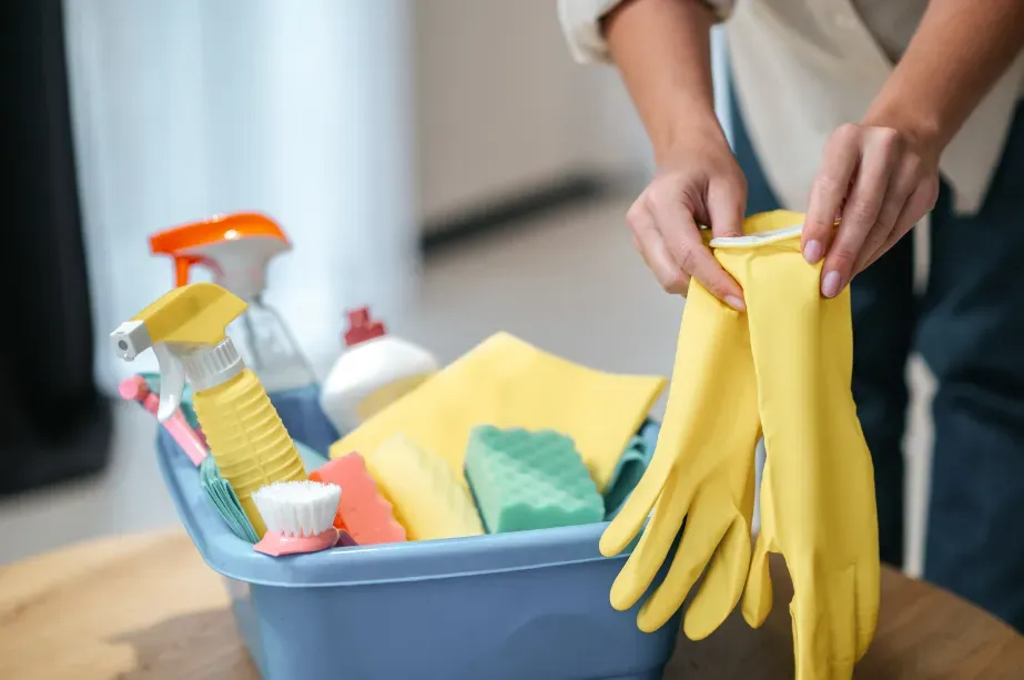 Hands putting on yellow rubber gloves; cleaning supplies in a blue bucket on a table.