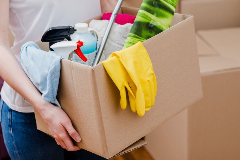 Woman holding a box filled with cleaning supplies, including spray bottles, gloves, and cloths.