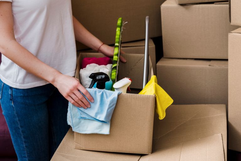 Person holding a box of cleaning supplies among moving boxes.