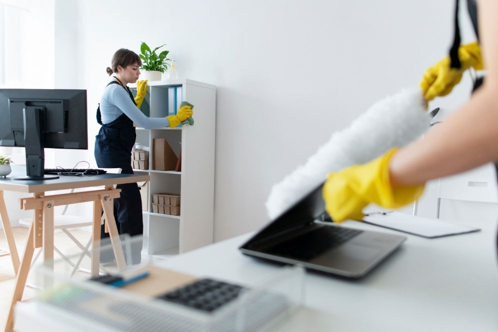 Two people cleaning an office: one dusting shelves, another dusting a laptop. Both wear yellow gloves.
