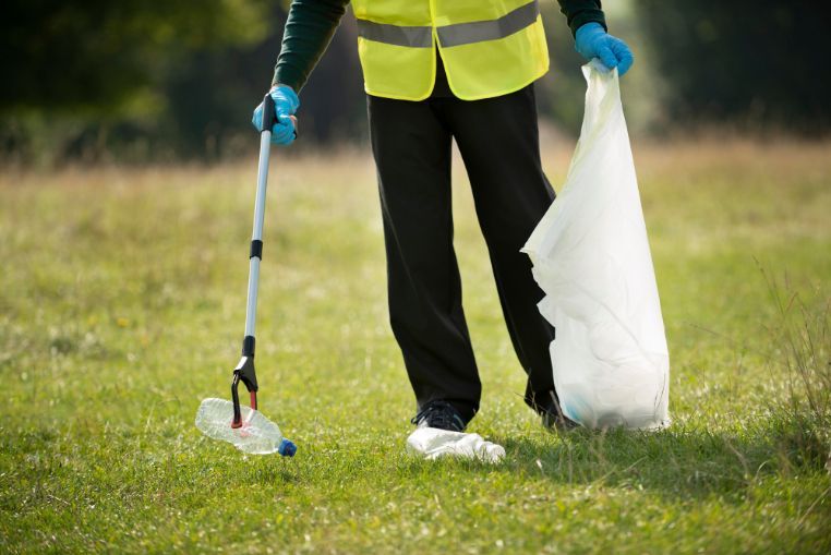 Person picking up trash in a grassy field with a grabber and a trash bag, wearing a yellow vest.