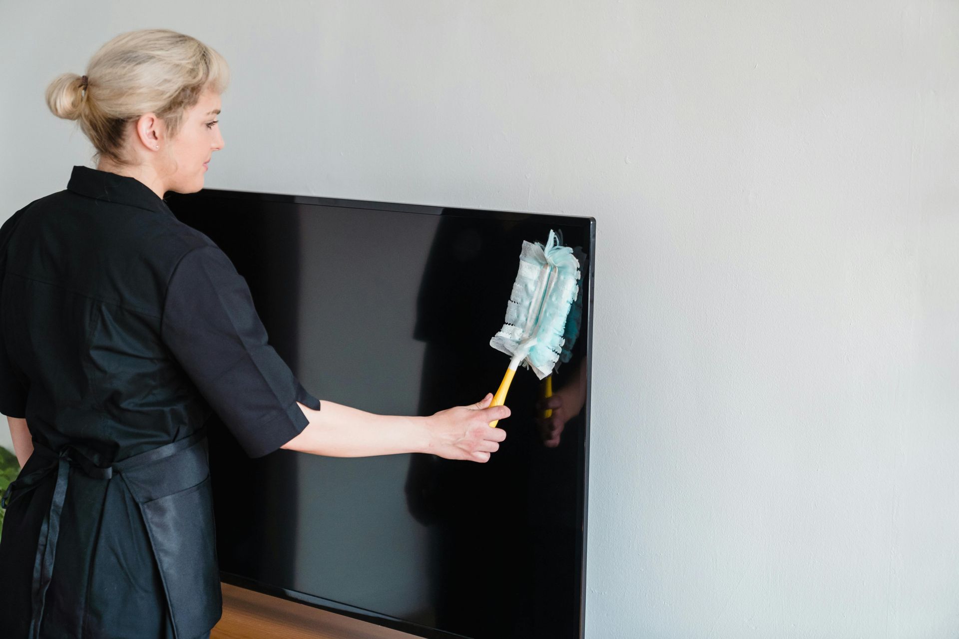 Woman dusting a large, black television with a yellow-handled duster; light-colored wall in the background.