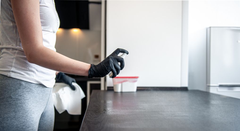 Person wearing black gloves sprays cleaning product on a kitchen counter, holding a wipe.