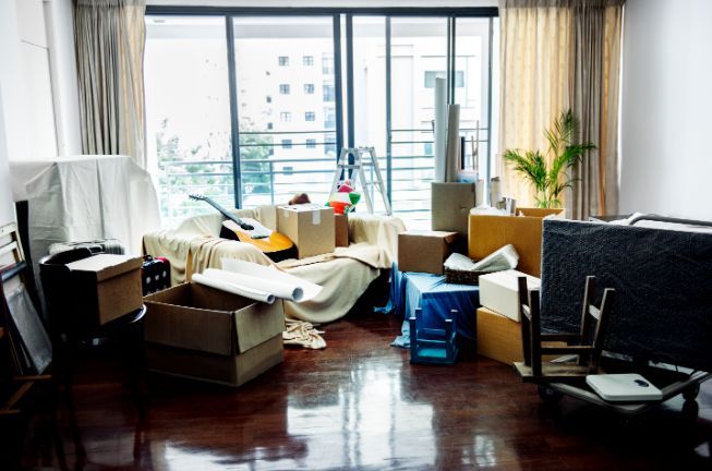 A cluttered room during a move, with packed boxes, furniture, and a guitar by a large window.