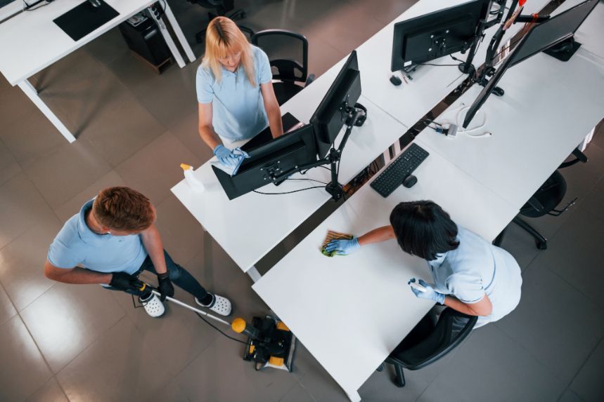 Three people cleaning an office: one vacuuming, two wiping desks. White desks, blue shirts.