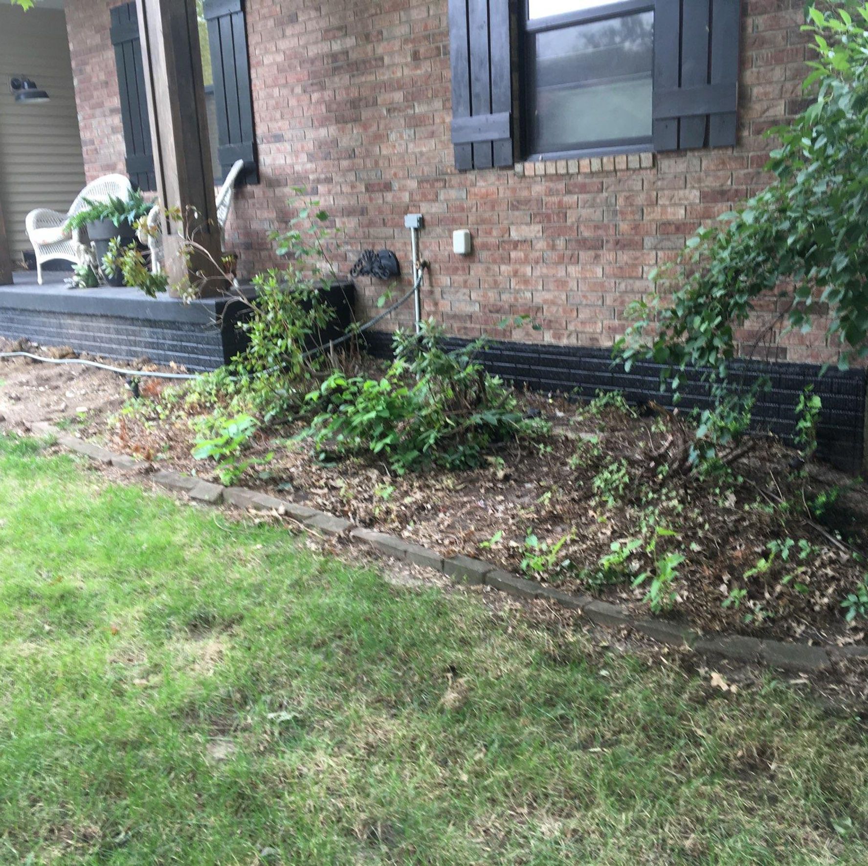 Brick house exterior with black shutters and a narrow garden bed along the foundation.