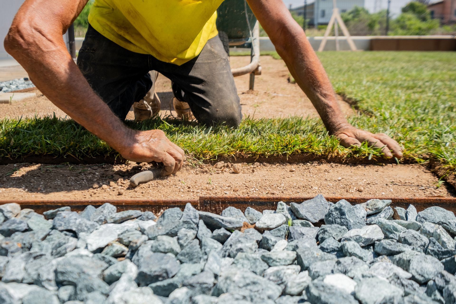 A person kneeling, laying sod in a yard with gravel and a wooden border.
