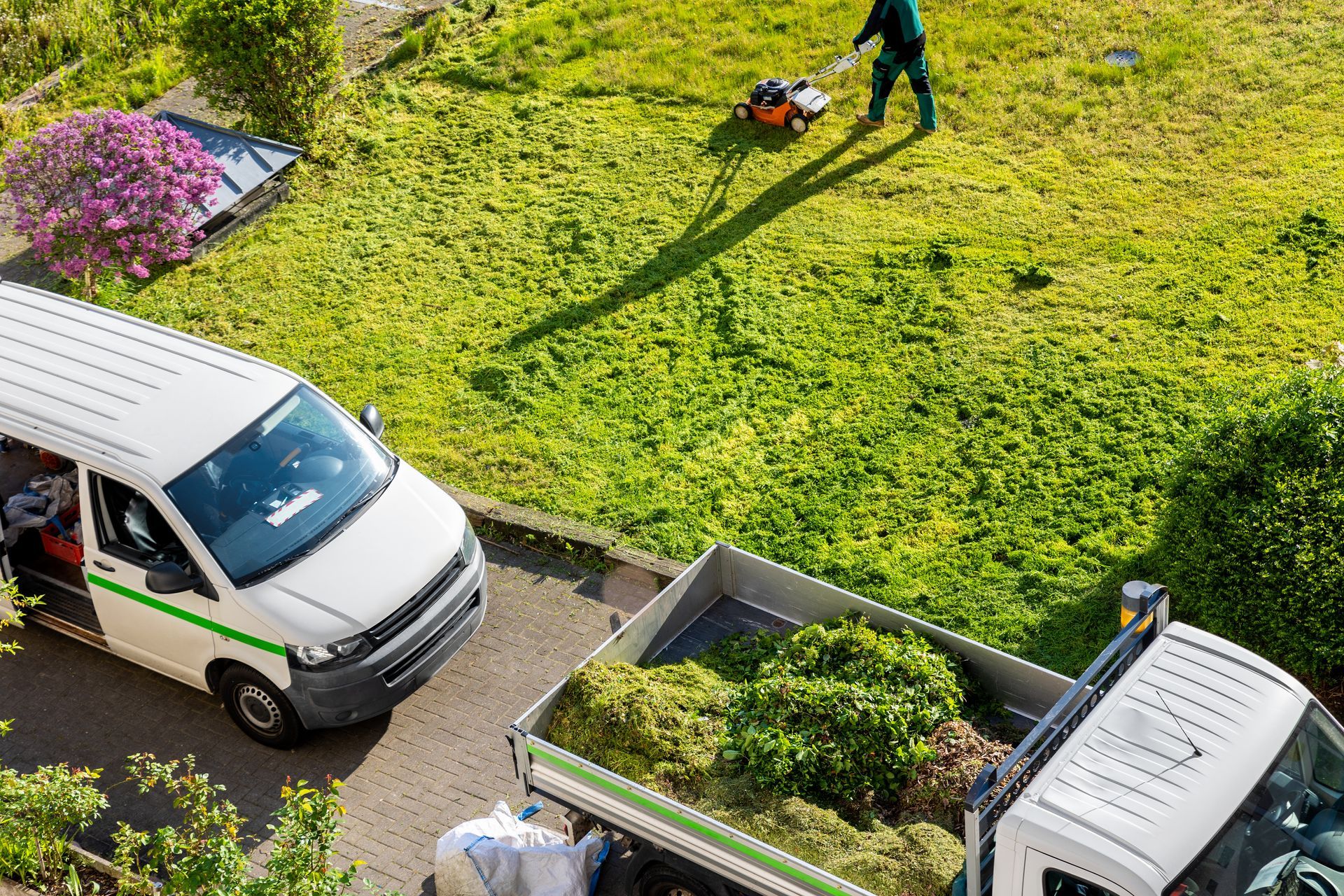 Lawn care service: worker mowing lawn; van and truck with yard waste on a sunny day.