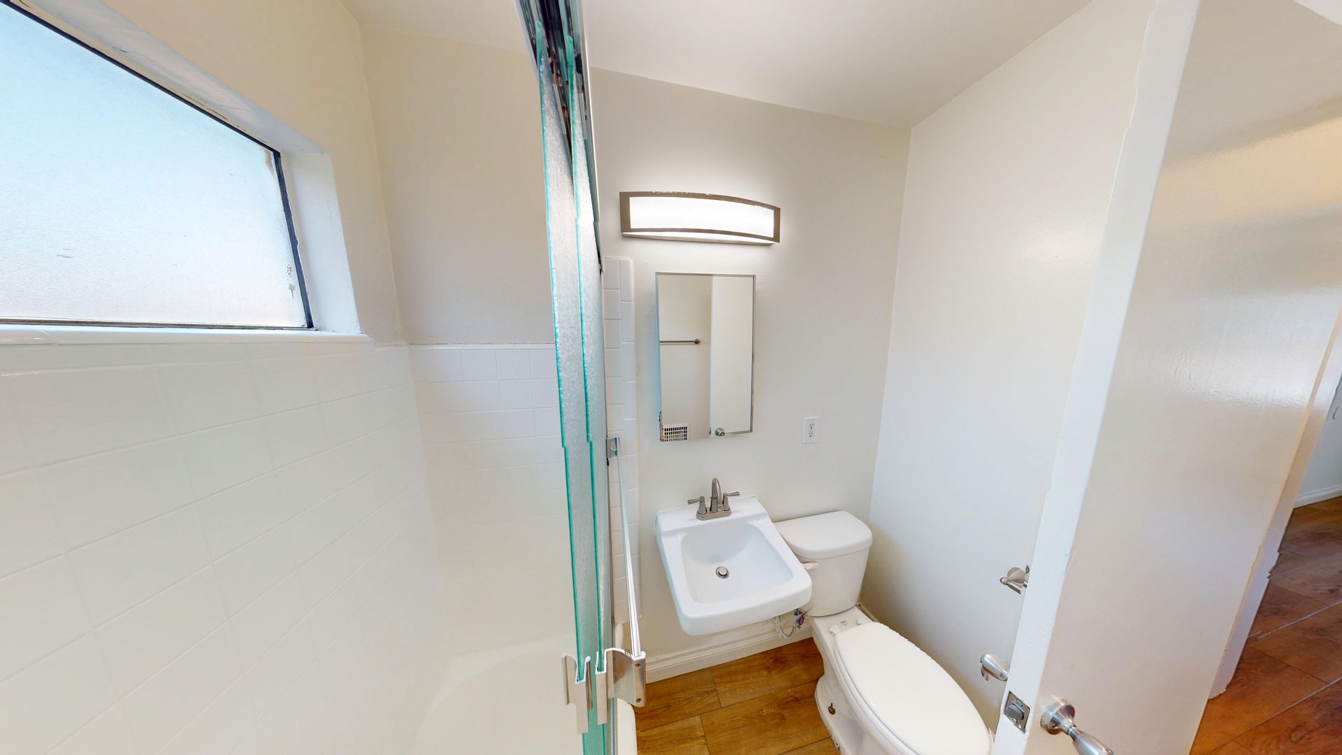 Bathroom with a sink, toilet, mirror, and shower. White walls and brown tiled floor.