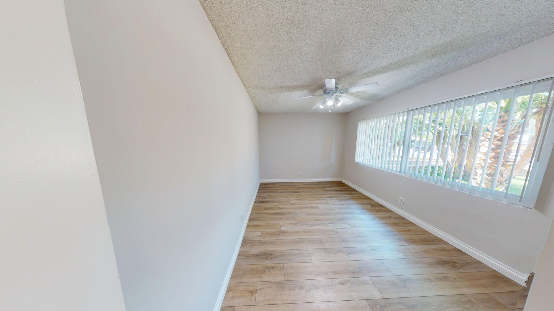 Empty room with light wood flooring, a window with blinds, and a ceiling light.