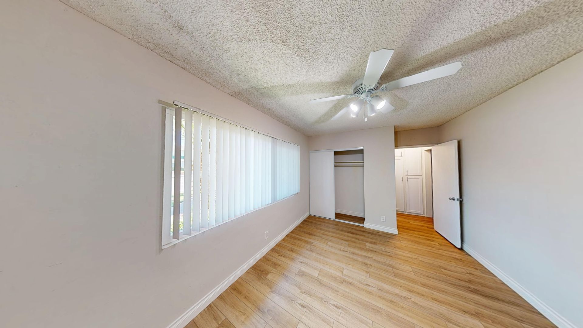 Empty bedroom with light wood floor, white walls, closed window with blinds, ceiling fan, and open closet.