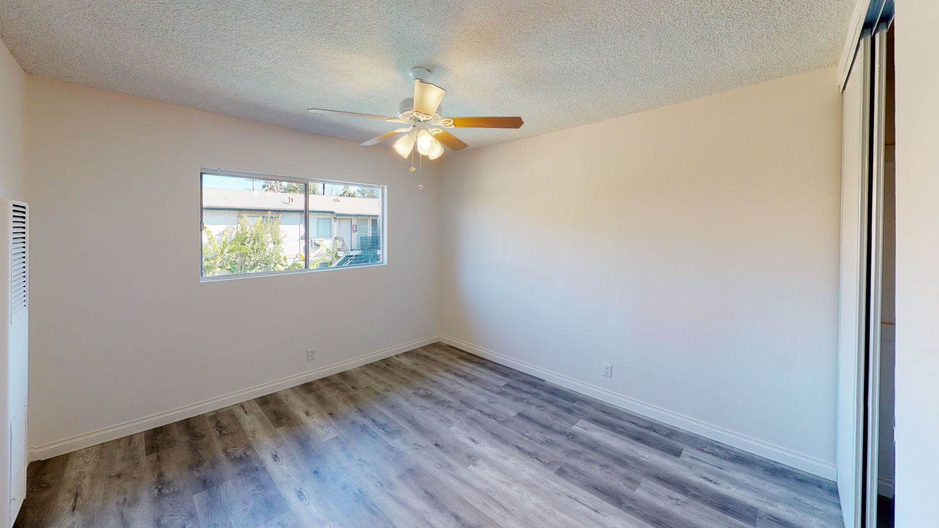 Empty room with a window, ceiling fan, and wood-look flooring. White walls and a sliding door.