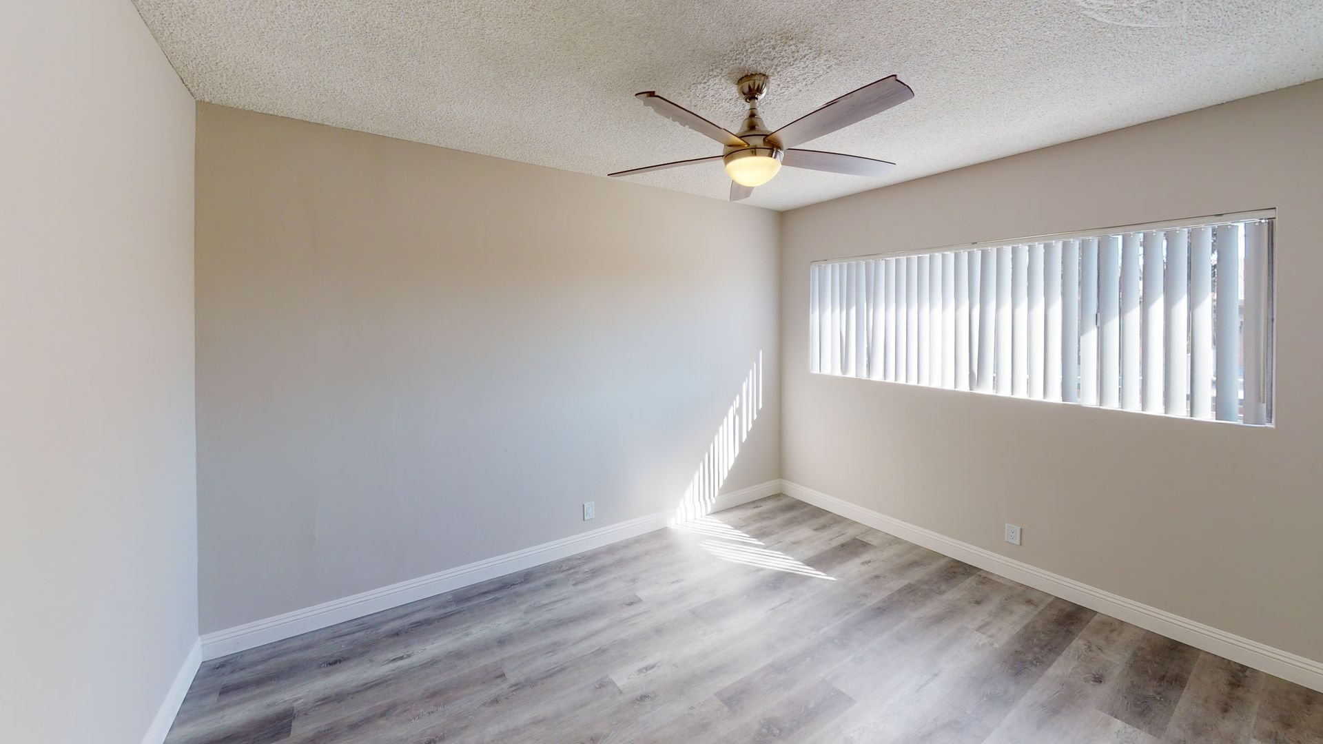 Empty bedroom with gray laminate flooring, beige walls, and a ceiling fan. A window with closed blinds allows sunlight.