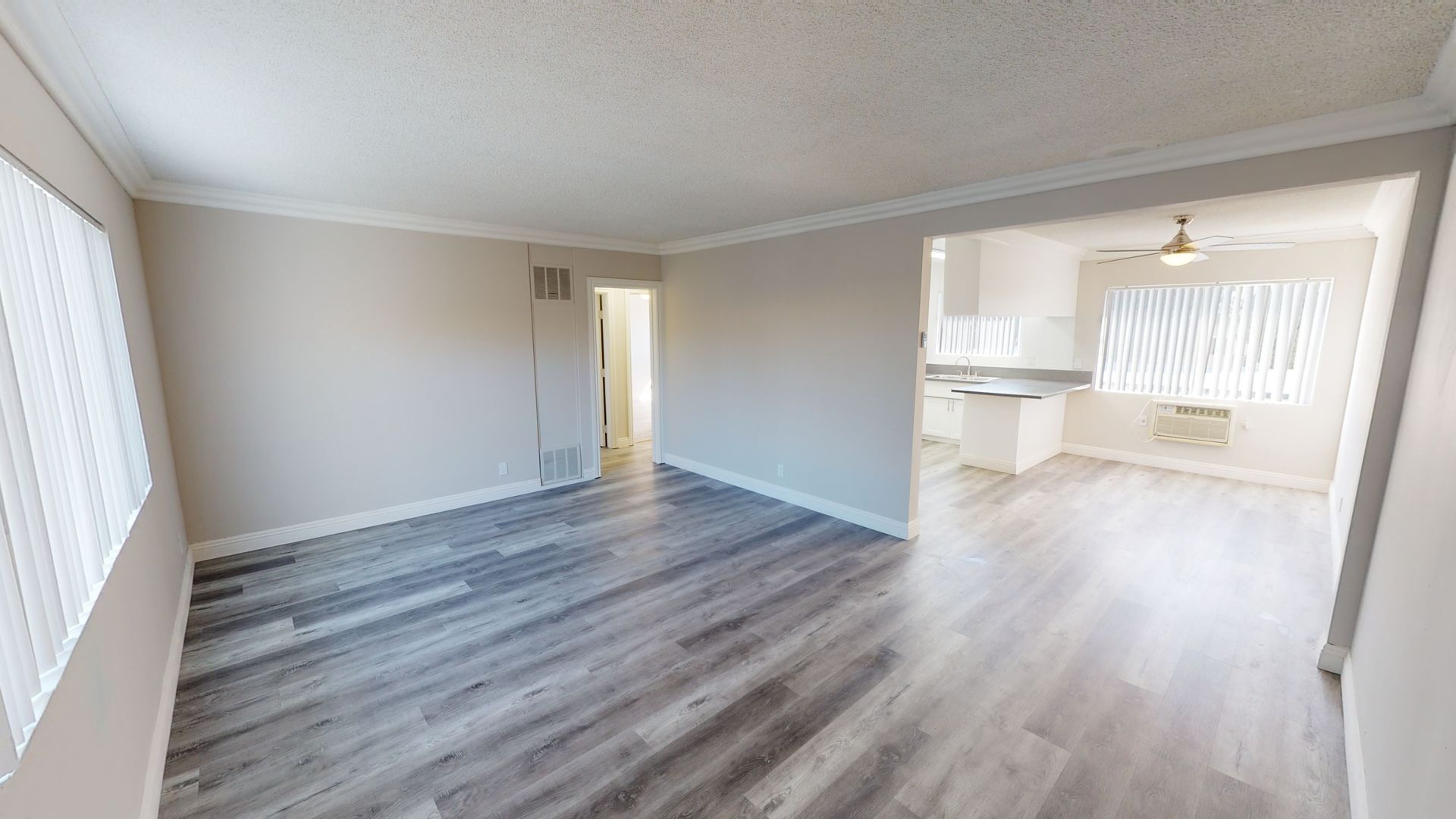 Interior view of a living space with grey wood flooring, white walls, and an adjacent kitchen.
