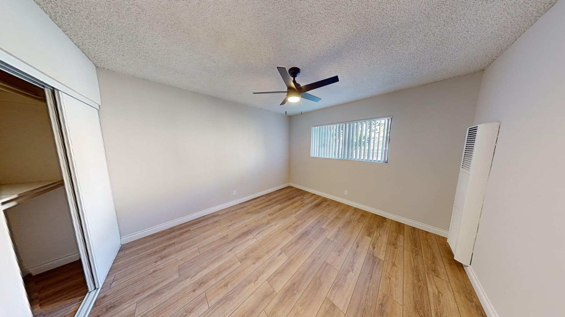 Empty bedroom with wood-look floors, closet, ceiling fan, window with blinds, and a white wall-mounted heater.