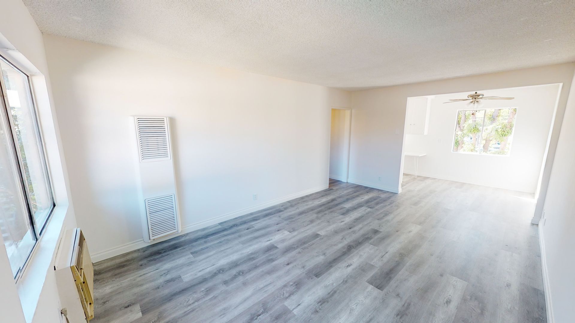 Empty living room with gray wood-look floor, white walls, window, and doorway to another room.