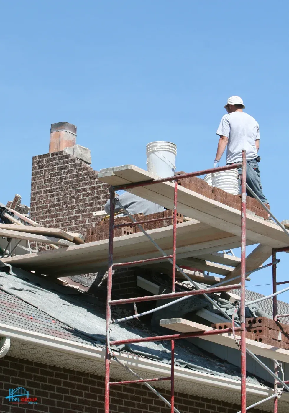 Worker on scaffold painting a brick chimney on a house roof under a blue sky