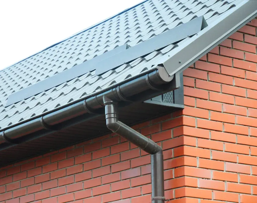 Brick house corner with gray metal roof and dark gutter downspout