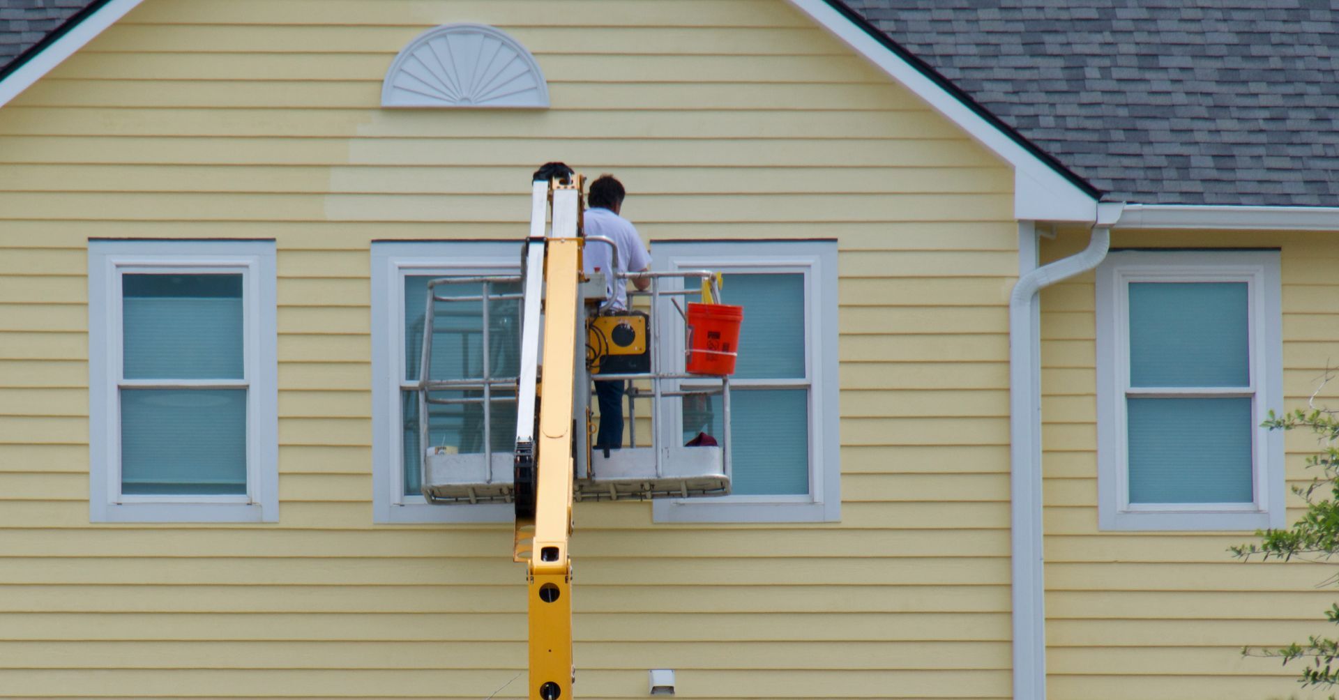 A man is painting a window on a yellow house.