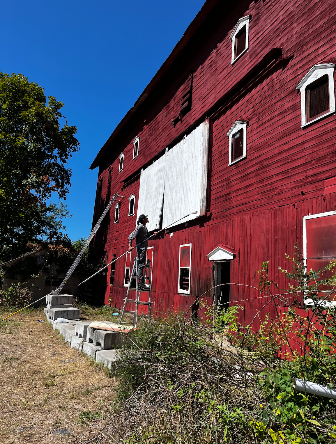 A man is painting the side of a red barn.