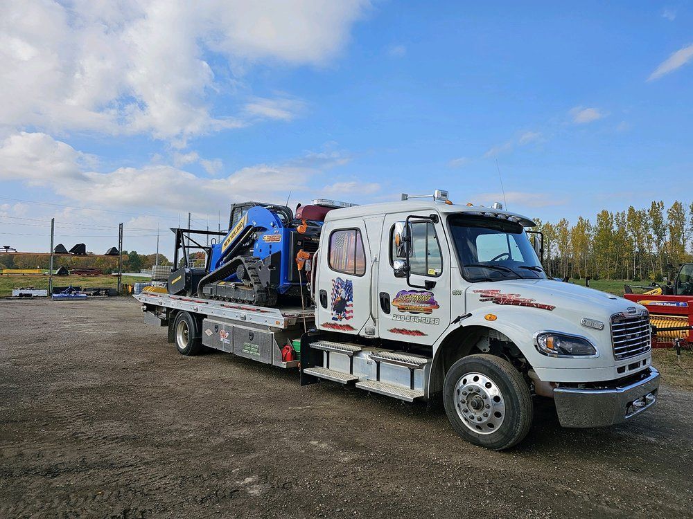 White tow truck transporting a blue excavator on a sunny day.