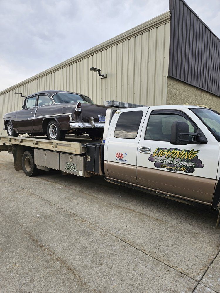 Brown classic car being towed on a flatbed tow truck with business name on its door in front of a building.