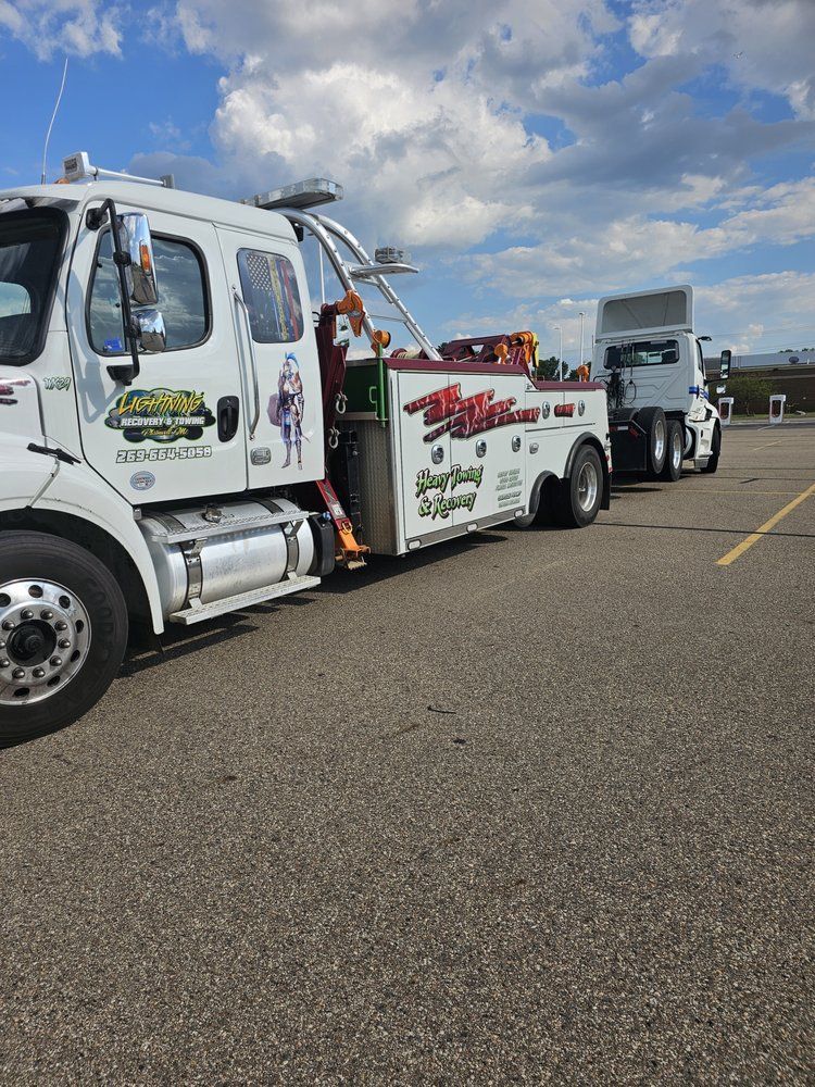 Tow truck towing a semi-truck on asphalt; clear sky. The tow truck is white with red and green details.
