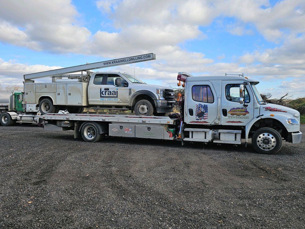 Tow truck carrying a white work truck on a flatbed, under a cloudy sky.