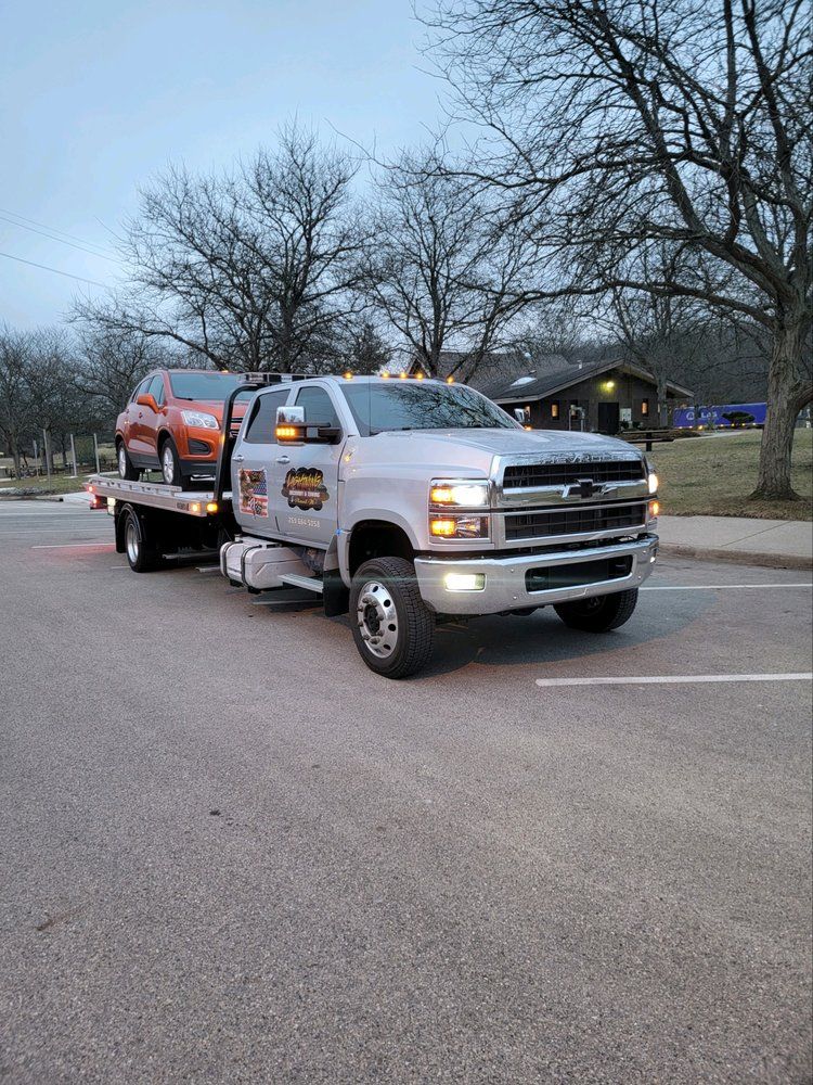 Silver tow truck with an orange car on its flatbed in a parking lot.